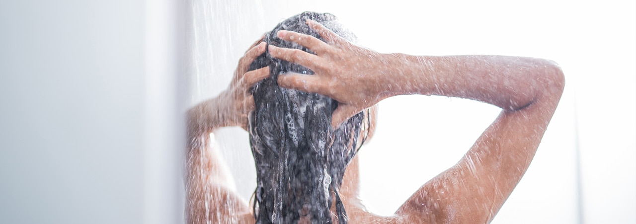 Mulher de costas no chuveiro, com as mãos na cabeça lavando os cabelos com um shampoo detox.