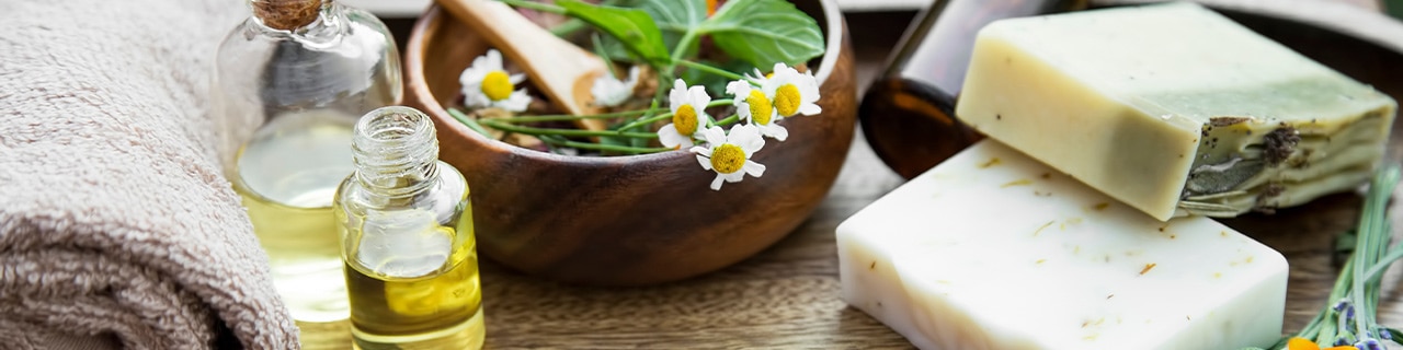 Sabonetes em barra, frascos com óleo essencial e toalha ao lado de ervas e flores amarelas em uma bancada de madeira.