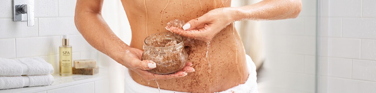 Pessoa realizando esfoliação corporal com esponja e creme esfoliante em pote de vidro, com a pele marcada por partículas e espuma, em ambiente de spa