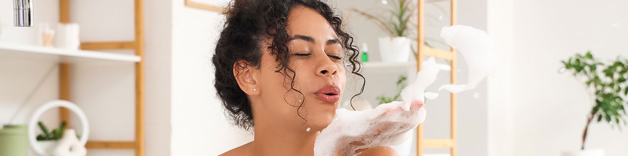 Mulher com cabelo cacheado no banheiro toma banho e ensaboa o corpo com espuma branca no chuveiro, com lavatório e plantas ao fundo.