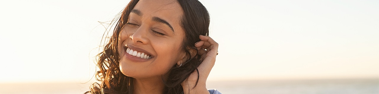 Mulher sorridente com cabelos soltos e protegidos por protetor solar capilar em uma praia ensolarada, simbolizando cuidados com o cabelo no verão.